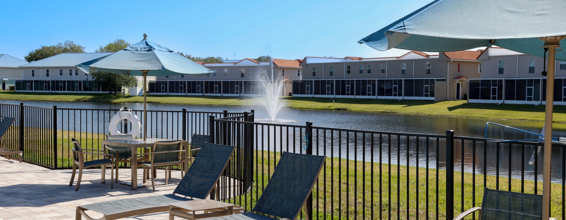 Poolside lounge area at Heritage Key Villas in Kissimmee, Florida, featuring modern patio furniture and umbrellas overlooking a scenic lake with a fountain. These pet-friendly and family-friendly apartments for rent offer luxury outdoor amenities for resi