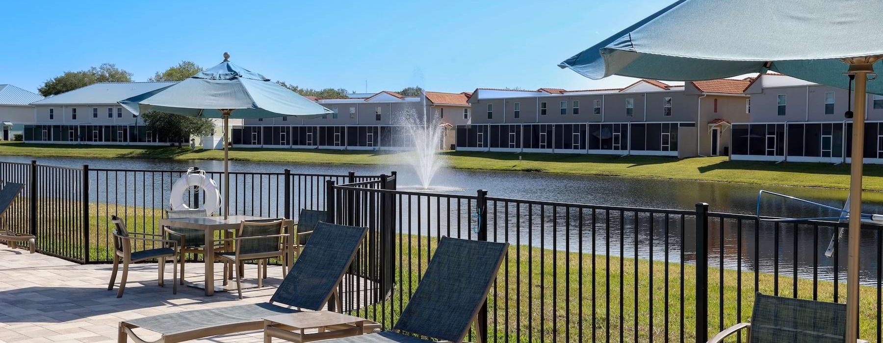 Poolside lounge area at Heritage Key Villas in Kissimmee, Florida, featuring modern patio furniture and umbrellas overlooking a scenic lake with a fountain. These pet-friendly and family-friendly apartments for rent offer luxury outdoor amenities for resi